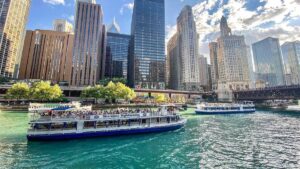 Wide shot of a boat doing a river tour with large city buildings behind it on a sunny day in Chicago, Illinois